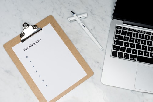 A blank packing list on a clipboard is placed on a marble surface beside a pen and an open laptop.