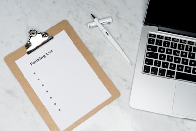 A blank packing list on a clipboard is placed on a marble surface beside a pen and an open laptop.