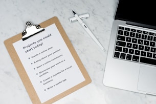 A marble surface displays a clipboard holding a paper with a list titled 'Projects you could start today!', placed next to an open laptop and a pen. The list suggests ideas like creating an online shop or a useful app.