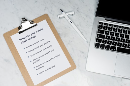 A marble surface displays a clipboard holding a paper with a list titled 'Projects you could start today!', placed next to an open laptop and a pen. The list suggests ideas like creating an online shop or a useful app.