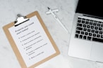 A marble surface displays a clipboard holding a paper with a list titled 'Projects you could start today!', placed next to an open laptop and a pen. The list suggests ideas like creating an online shop or a useful app.