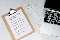 A marble surface displays a clipboard holding a paper with a list titled 'Projects you could start today!', placed next to an open laptop and a pen. The list suggests ideas like creating an online shop or a useful app.