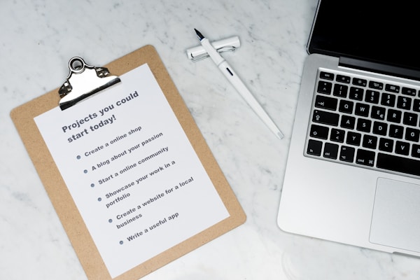 A marble surface displays a clipboard holding a paper with a list titled 'Projects you could start today!', placed next to an open laptop and a pen. The list suggests ideas like creating an online shop or a useful app.
