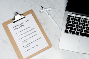 A marble surface displays a clipboard holding a paper with a list titled 'Projects you could start today!', placed next to an open laptop and a pen. The list suggests ideas like creating an online shop or a useful app.
