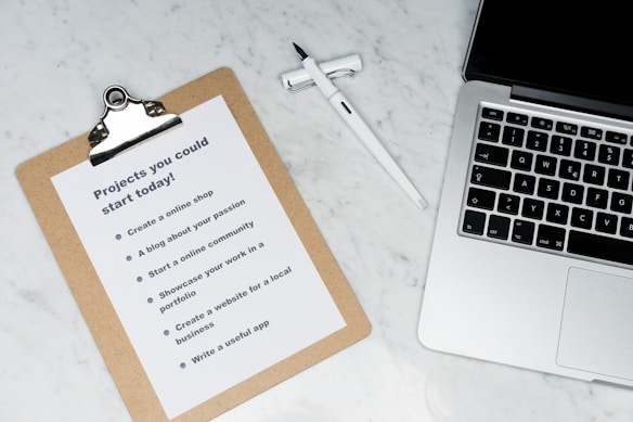 A marble surface displays a clipboard holding a paper with a list titled 'Projects you could start today!', placed next to an open laptop and a pen. The list suggests ideas like creating an online shop or a useful app.