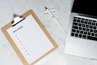 A marble surface holds a silver laptop partially visible on the right. To the left, there is a clipboard with a blank list titled 'Date Night'. A pen rests between the laptop and the clipboard.