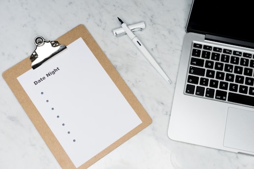 A marble surface holds a silver laptop partially visible on the right. To the left, there is a clipboard with a blank list titled 'Date Night'. A pen rests between the laptop and the clipboard.