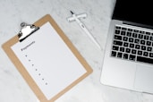 A clipboard with a blank payments checklist is placed on a marble surface next to an open laptop and a white pen. The setup appears to be organized for financial planning or administrative work.