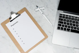 A clipboard with a blank payments checklist is placed on a marble surface next to an open laptop and a white pen. The setup appears to be organized for financial planning or administrative work.