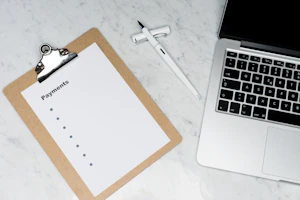 A clipboard with a blank payments checklist is placed on a marble surface next to an open laptop and a white pen. The setup appears to be organized for financial planning or administrative work.