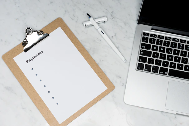 A clipboard with a blank payments checklist is placed on a marble surface next to an open laptop and a white pen. The setup appears to be organized for financial planning or administrative work.