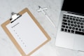 A clipboard with a blank payments checklist is placed on a marble surface next to an open laptop and a white pen. The setup appears to be organized for financial planning or administrative work.