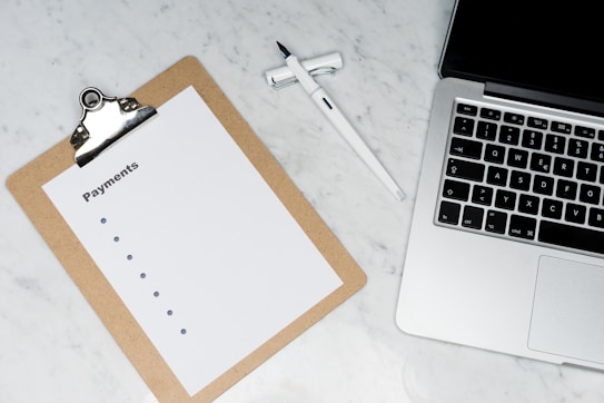 A clipboard with a blank payments checklist is placed on a marble surface next to an open laptop and a white pen. The setup appears to be organized for financial planning or administrative work.