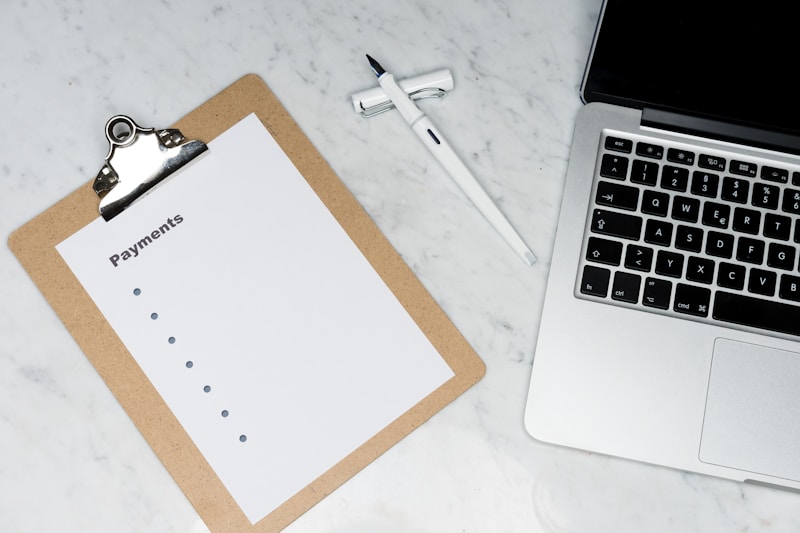 A clipboard with a blank payments checklist is placed on a marble surface next to an open laptop and a white pen. The setup appears to be organized for financial planning or administrative work.