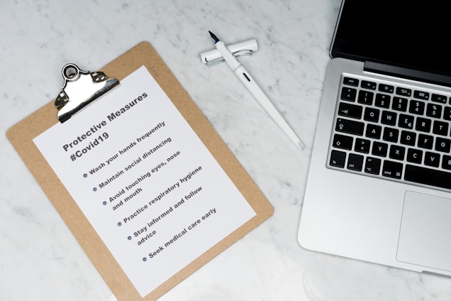 A clipboard with a paper titled 'Protective Measures #Covid19' lists safety guidelines. It includes steps such as washing hands, maintaining social distance, and seeking medical care early. Positioned on a marble surface, next to a silver laptop and a white pen.