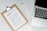 A clipboard with a paper titled 'Protective Measures #Covid19' lists safety guidelines. It includes steps such as washing hands, maintaining social distance, and seeking medical care early. Positioned on a marble surface, next to a silver laptop and a white pen.