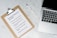 A clipboard with a paper titled 'Protective Measures #Covid19' lists safety guidelines. It includes steps such as washing hands, maintaining social distance, and seeking medical care early. Positioned on a marble surface, next to a silver laptop and a white pen.