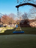 A playground swing surrounded by caution tape, situated in an open grassy area. The swing appears unused and is secured with chains, casting a shadow on the ground. Trees in the background display bare or sparsely leaved branches, suggesting a late autumn or early winter setting. A fence and some buildings are visible in the distance under a clear blue sky.