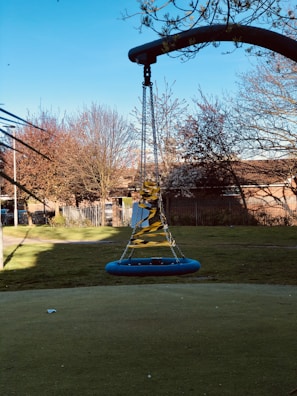 A playground swing surrounded by caution tape, situated in an open grassy area. The swing appears unused and is secured with chains, casting a shadow on the ground. Trees in the background display bare or sparsely leaved branches, suggesting a late autumn or early winter setting. A fence and some buildings are visible in the distance under a clear blue sky.