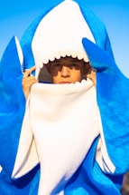 A person is dressed in a blue and white shark costume, with a hood shaped like a shark's open mouth. The background is a clear blue sky.