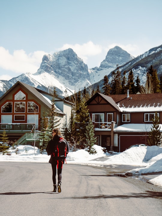 person in red jacket standing on snow covered ground near brown and white house during daytime in The Three Sisters Canada