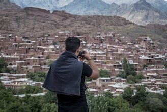 A person with a dark shirt and a towel draped over the shoulders is taking a photo of a village. The village consists of numerous brick houses tightly packed together on a hillside. Green trees are scattered throughout, and jagged mountains rise in the background, partially covered by clouds.