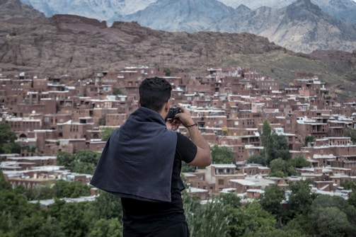 A person with a dark shirt and a towel draped over the shoulders is taking a photo of a village. The village consists of numerous brick houses tightly packed together on a hillside. Green trees are scattered throughout, and jagged mountains rise in the background, partially covered by clouds.