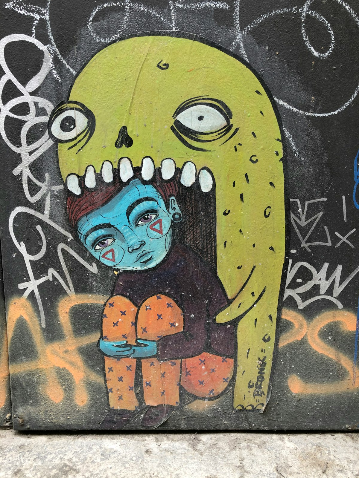 Graffit style drawing.  There is a like green monster with one dilated pupil and one regular pupil.  It has a huge mouth and fearsome teeth.  In his mouth is a blue skinned boy wearing a brown sweater and orange pants with little x's. He has two red inverted triangles on his cheeks and seems sad.