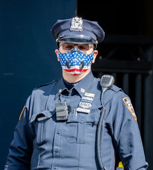 A person is dressed in a police officer's uniform, wearing a cap, sunglasses, and a face mask decorated with the American flag. The uniform has badges and a badge number visible. A radio is attached to the shoulder, with a cable running down the front.