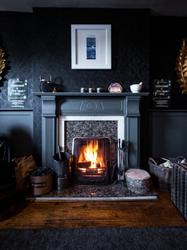 A cozy fireplace in a dark, elegantly decorated room. The fireplace mantle is adorned with small objects and framed art is hung above it. A log is burning brightly in the fireplace, creating a warm and inviting atmosphere. Various items such as a basket, logs, and fireplace tools are arranged neatly around the hearth, contributing to the homely feel.