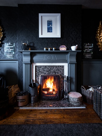 A cozy fireplace in a dark, elegantly decorated room. The fireplace mantle is adorned with small objects and framed art is hung above it. A log is burning brightly in the fireplace, creating a warm and inviting atmosphere. Various items such as a basket, logs, and fireplace tools are arranged neatly around the hearth, contributing to the homely feel.