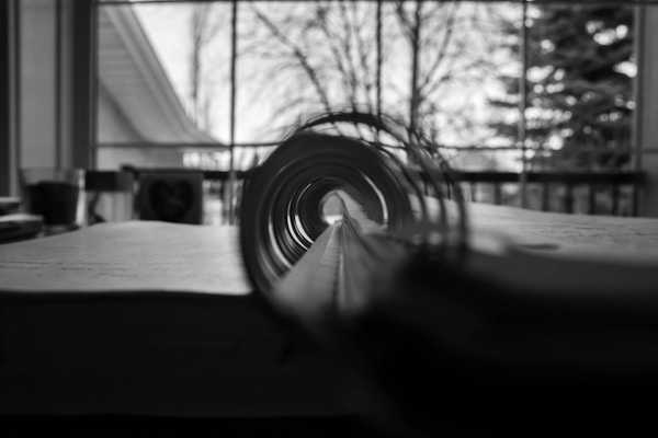A black and white photo capturing the spiral binding of a notebook in sharp focus, creating a tunnel-like effect. The background is blurred, showing trees and a building through a window.