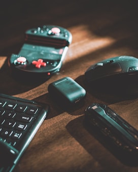 A collection of tech gadgets resting on a wooden surface includes a gaming controller with red buttons, a wireless mouse, a closed pair of wireless earbuds in their case, a keyboard, and a USB flash drive.