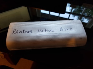 A white oblong container with horizontal ridges on its surface has the words 'Random Useful Bits' handwritten on it in black ink. The container is placed on a dark, reflective surface near a window, allowing natural light to illuminate the scene.