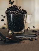 A transparent glass mug overflowing with dark roasted coffee beans is placed on top of a closed notebook. Some coffee beans are scattered around and appear to be in motion, emphasizing a sense of dynamic stillness. The scene is softly lit, casting gentle shadows.