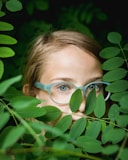 girl in blue framed eyeglasses hiding behind green leaves