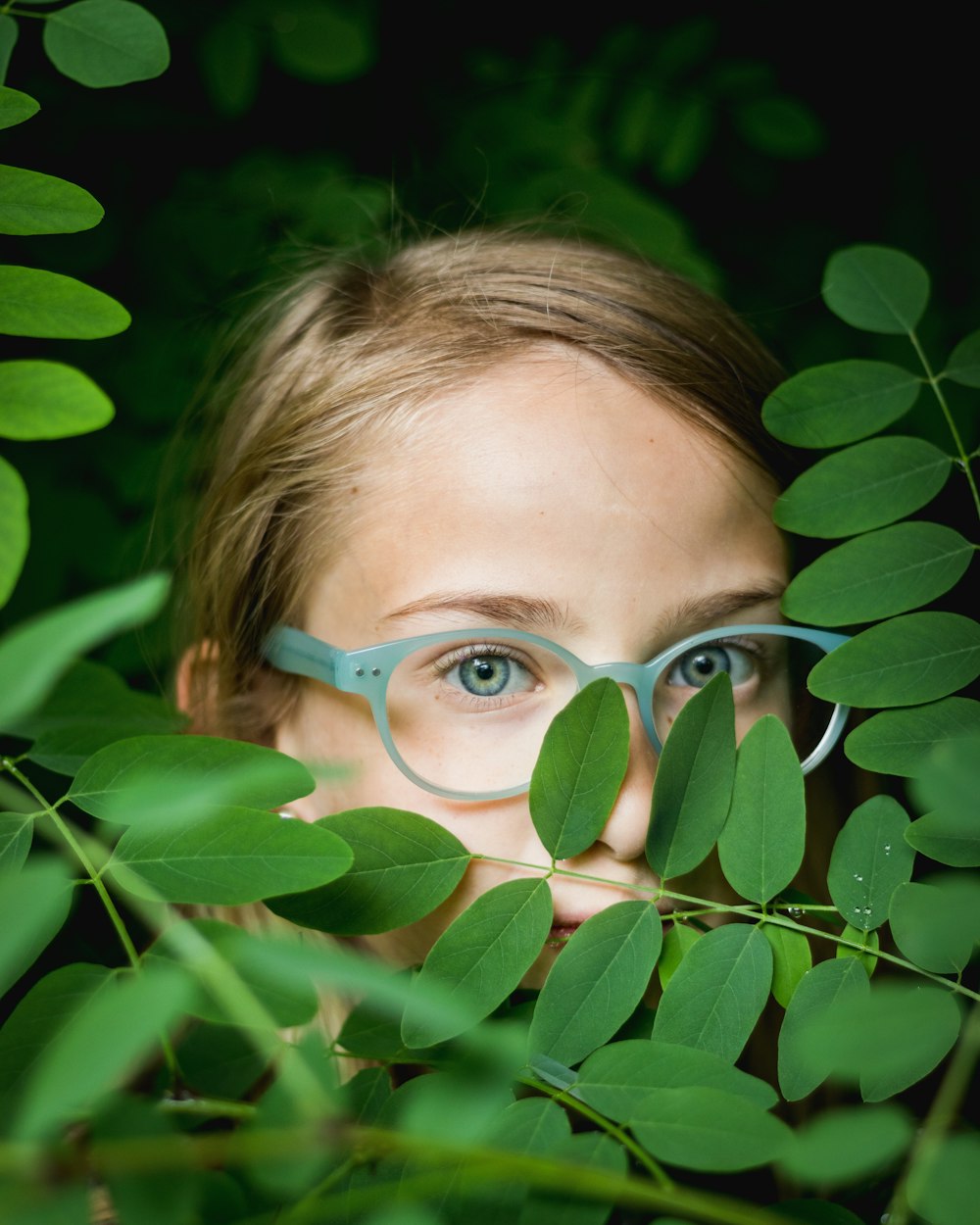 緑の葉の後ろに隠れている青いフレームの眼鏡の女の子
