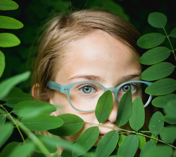 girl in blue framed eyeglasses hiding behind green leaves