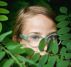 girl in blue framed eyeglasses hiding behind green leaves