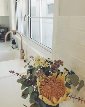 A kitchen sink area with a gold-colored faucet is complemented by a bouquet of flowers on the counter. The window behind the sink allows natural light to brighten the white-tiled backsplash and countertop. A soap dispenser is visible next to the faucet.