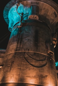 A large structure with a cylindrical shape and intricate details, featuring wires and cables attached to its surface. The material appears to be metallic or concrete, illuminated by warm and cool lighting that creates an industrial, futuristic ambiance.