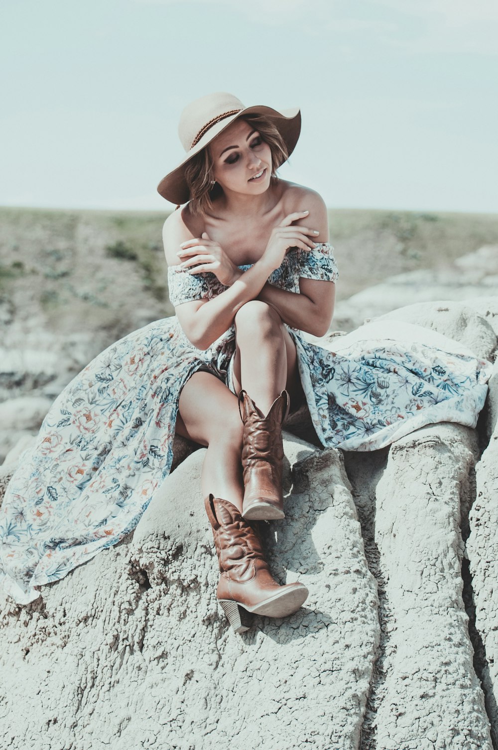 Mujer en vestido floral azul y blanco con botas de vaquero marrones sentada en roca gris durante