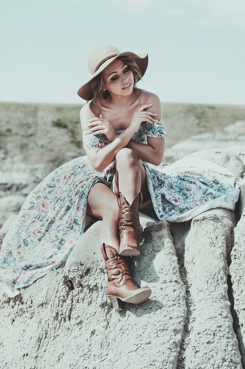 woman in blue and white floral dress wearing brown cowboy boots sitting on gray rock during