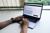 A person is typing on a laptop situated on a clean, light-colored desk. The screen displays text with some highlighted sections, suggesting work or study. The hands are positioned on the keyboard, and the person is wearing a watch.