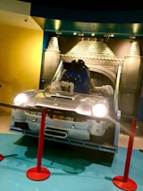 A large blue M&M character, wearing sunglasses, is seated inside a silver car resembling a Mini Cooper. The background features a decorative wall that resembles a castle facade. Red stanchion ropes are positioned around the display, which seems to be an exhibit or installation in a public space. Lights shine onto the scene, and the area is well-lit with a combination of cool and warm tones.