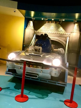 A large blue M&M character, wearing sunglasses, is seated inside a silver car resembling a Mini Cooper. The background features a decorative wall that resembles a castle facade. Red stanchion ropes are positioned around the display, which seems to be an exhibit or installation in a public space. Lights shine onto the scene, and the area is well-lit with a combination of cool and warm tones.
