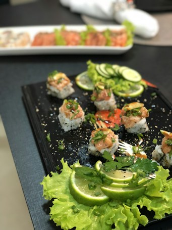 A black plate is artfully arranged with sushi rolls topped with salmon and chopped green onions. Surrounding the rolls are garnishings of sliced limes, fresh lettuce, and cucumber slices. Sesame seeds are scattered on the sushi rolls, adding texture. In the background, another plate with more sushi rolls can be slightly seen, indicating a culinary presentation setting.