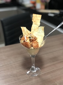 A glass goblet is filled with a culinary dish, featuring chunks of golden-brown crispy chicken topped with garnish, accompanied by large, thin pieces of seasoned flatbread. A spoon is placed in the goblet, and it rests on a wooden dining surface with a blurred background.