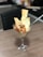 A glass goblet is filled with a culinary dish, featuring chunks of golden-brown crispy chicken topped with garnish, accompanied by large, thin pieces of seasoned flatbread. A spoon is placed in the goblet, and it rests on a wooden dining surface with a blurred background.