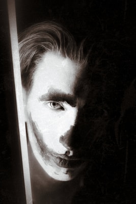 A highly stylized, monochrome image features a person's face partially illuminated with dramatic lighting. The face is painted with white makeup and dark shadows, creating a stark contrast. The light source is on the left side, highlighting one eye and casting shadow on the rest of the face.